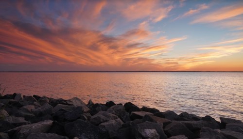 A serene sunset over a calm lake, with vibrant orange and pink clouds reflecting on the water and rocky shoreline.