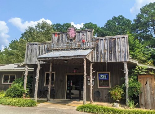 Exterior of Blind Pig BBQ, a rustic wooden building with a sign, surrounded by trees and a clear blue sky.