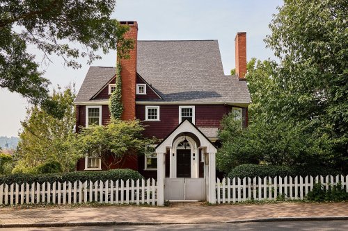 A charming red house with a gray roof, surrounded by greenery and a white picket fence.