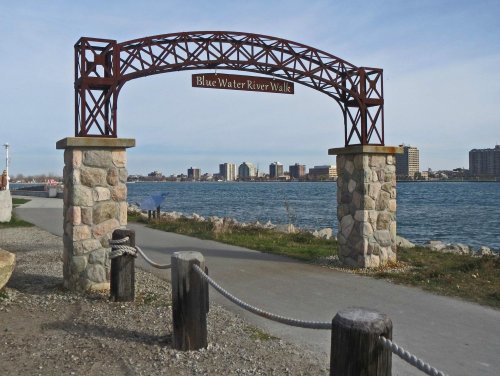 Archway sign for "Blue Water River Walk" with a waterfront view and city skyline in the background.