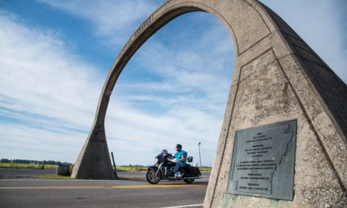 A motorcyclist rides under a large concrete arch with a plaque beside a rural road.