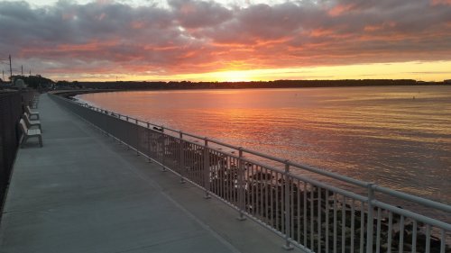 A serene waterfront path at sunset, with vibrant orange and pink hues reflecting on the calm water.