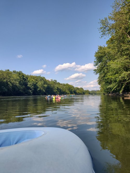 A serene river scene with kayakers paddling in the distance, surrounded by lush green trees and a clear blue sky.