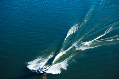 Aerial view of a boat towing a person on a wakeboard across calm water, creating wake trails behind.