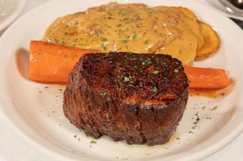A plate featuring a grilled steak, a large carrot, and a creamy sauce over sliced potatoes, garnished with herbs.