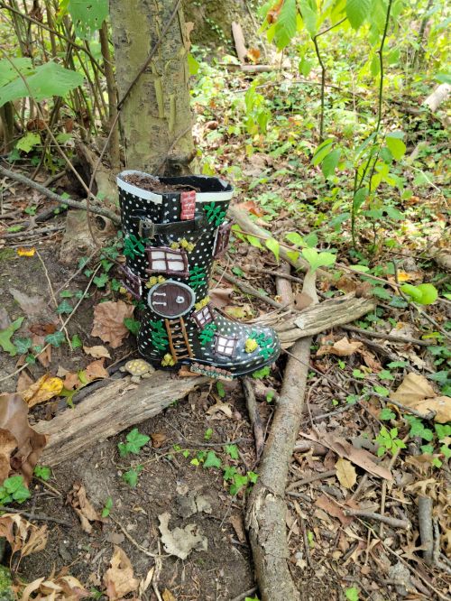 A colorful, decorated rubber boot stands among green foliage and fallen leaves in a wooded area.
