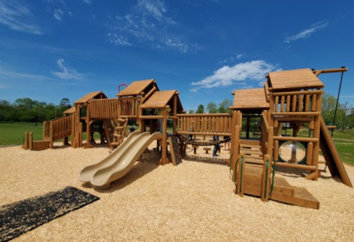 A wooden playground with slides, climbing structures, and swings, set against a clear blue sky and green grass.