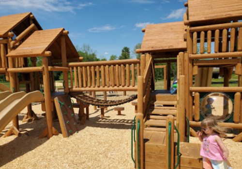 A wooden playground with slides, a bridge, and climbing features, set in a sunny outdoor park. A child is playing nearby.