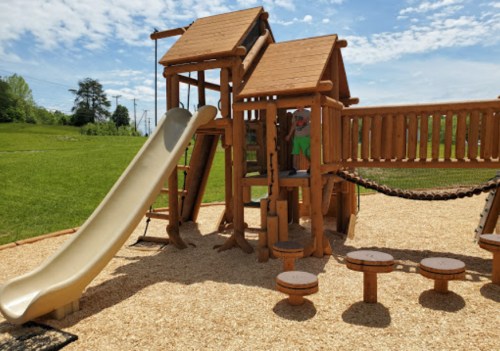 A wooden playground with a slide, climbing structure, and play area on a sunny day.