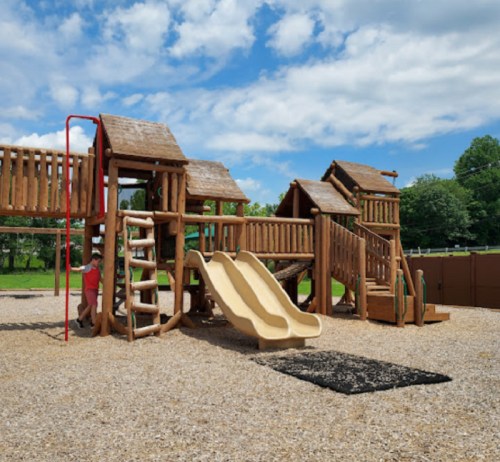 A wooden playground with slides, climbing structures, and a sandy area under a blue sky with clouds.