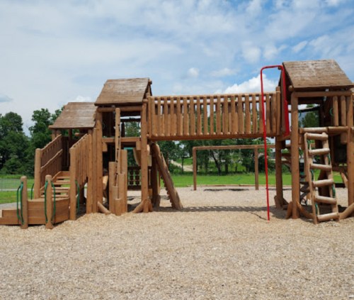 A wooden playground structure with slides, climbing features, and a bridge, set on a gravel surface under a blue sky.