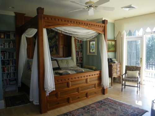 A cozy bedroom featuring a wooden four-poster bed with draped curtains, bookshelves, and a sunny window view.