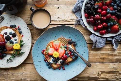 A rustic table set with a plate of toast topped with yogurt and berries, a cup of coffee, and a bowl of mixed fruits.