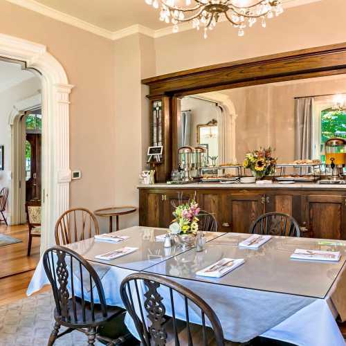 A bright dining room with a chandelier, wooden furniture, and a table set for a meal, surrounded by large windows.