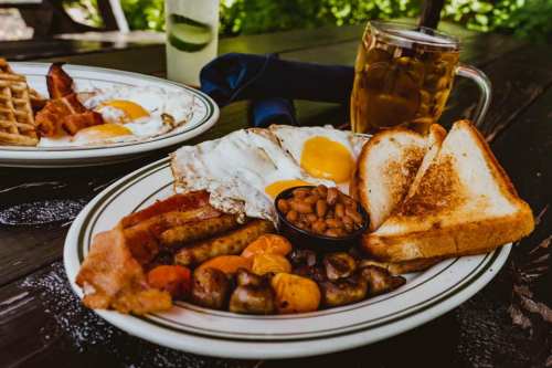A hearty breakfast plate with eggs, bacon, sausage, toast, baked beans, and mushrooms, alongside a glass of iced tea.
