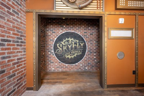 A brick wall with a circular logo reading "SkyFly Sugar America," framed by metal accents in a warm-toned room.