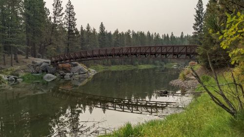 A wooden bridge spans a calm river, surrounded by trees and rocky banks, reflecting in the water.