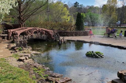 A scenic park with a stone bridge, pond, and children playing near a fountain surrounded by greenery.