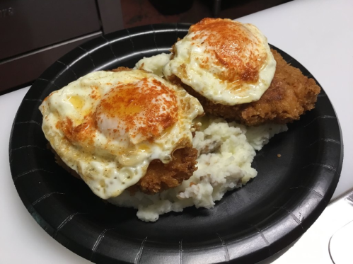 A black plate with two fried chicken pieces topped with sunny-side-up eggs, served over a bed of mashed potatoes.