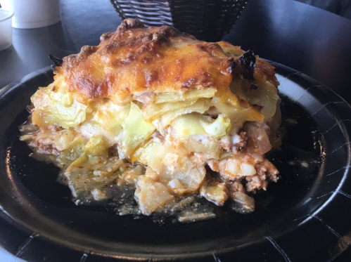 A slice of cheesy casserole with layers of meat, vegetables, and a golden-brown crust on a black plate.