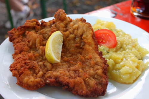 A plate featuring a large, crispy fried cutlet with a lemon wedge, served alongside creamy potato salad and a slice of tomato.