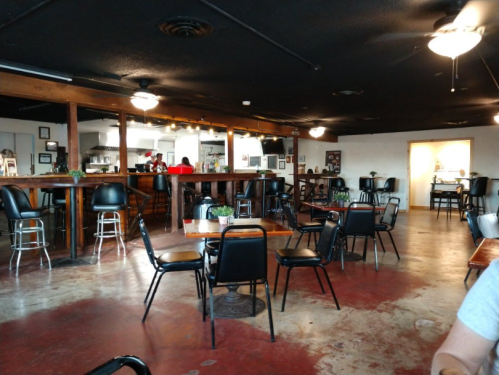 Interior of a casual restaurant with empty tables, black chairs, and a bar area in the background. Ceiling fans are visible.