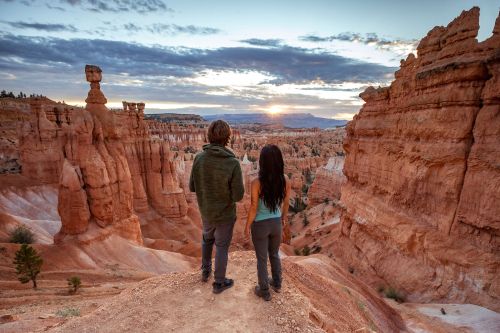 A couple stands on a rocky ledge, gazing at a stunning sunset over a canyon with unique rock formations.