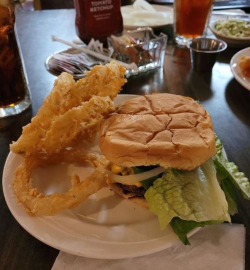 A plate with a burger topped with lettuce and cheese, accompanied by crispy onion rings and a drink in the background.