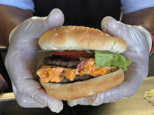 A person in gloves holds a burger with a sesame seed bun, lettuce, tomato, and a spicy orange sauce.