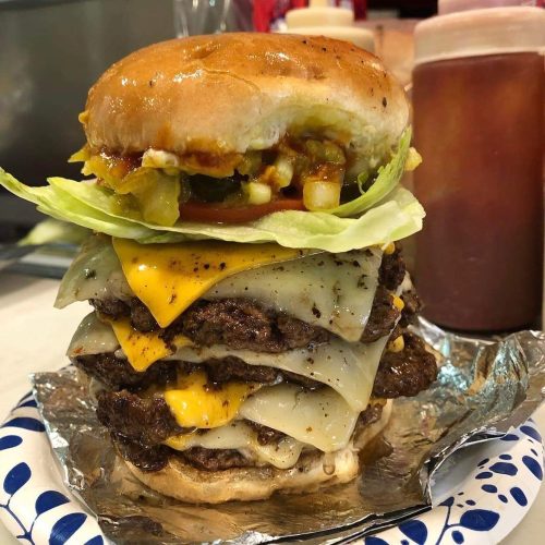 A towering burger with multiple beef patties, melted cheese, lettuce, and toppings, served on a paper plate.