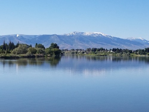 A serene lake reflects snow-capped mountains under a clear blue sky, surrounded by lush greenery.
