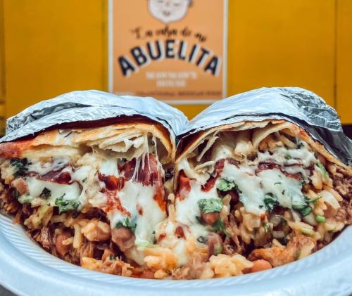 A close-up of a foil-wrapped burrito, filled with rice, meat, cheese, and garnished with herbs, on a white plate.