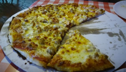 A partially eaten pizza with a golden crust, topped with cheese and various meats, on a checkered tablecloth.