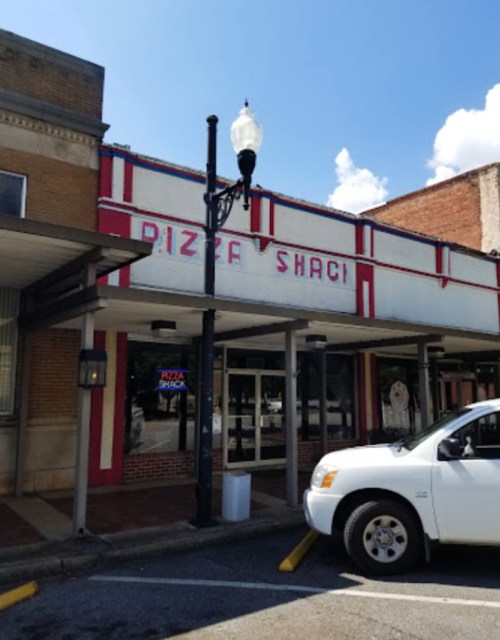 Exterior of a pizza restaurant called "Pizza Shack," featuring a bright sign and a parked white vehicle.