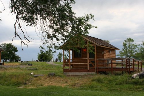 A small wooden cabin with a porch, surrounded by grassy fields and trees under a cloudy sky.
