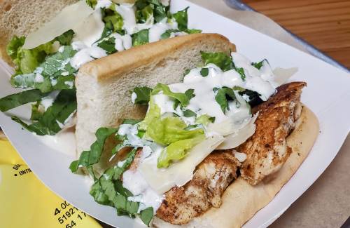 A sandwich with grilled chicken, lettuce, and creamy dressing on a soft bun, served in a takeout container.