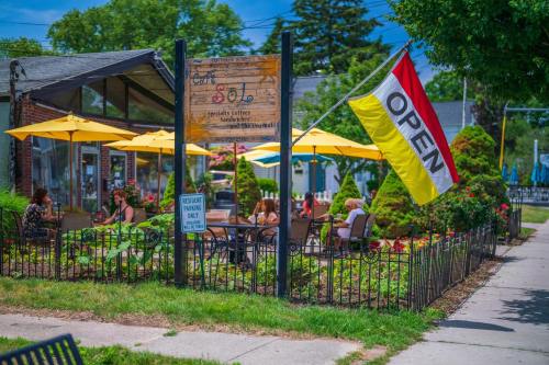 A cozy outdoor café with yellow umbrellas, a colorful "OPEN" flag, and patrons enjoying their meals in a garden setting.