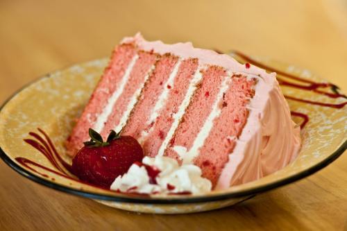 A slice of pink cake with layers, topped with frosting, a strawberry, and whipped cream on a decorative plate.
