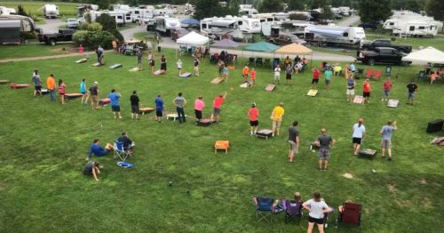 Aerial view of a lively outdoor event with people playing cornhole on a grassy field surrounded by tents and RVs.
