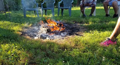 A campfire burns in a grassy area, surrounded by chairs and people sitting nearby, enjoying the outdoor setting.