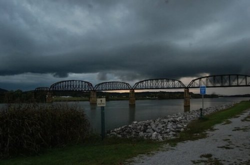 A dark, cloudy sky looms over a bridge spanning a river, with rocky banks and grassy areas nearby.