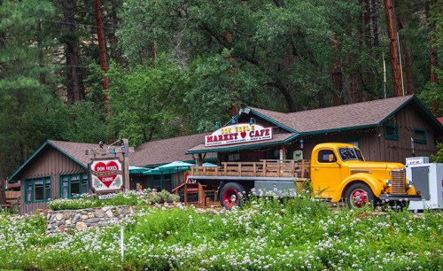 A rustic market and café with a yellow vintage truck parked outside, surrounded by greenery and wildflowers.