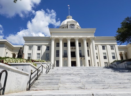 A grand white building with a dome and columns, featuring a statue in front and a blue sky with clouds above.