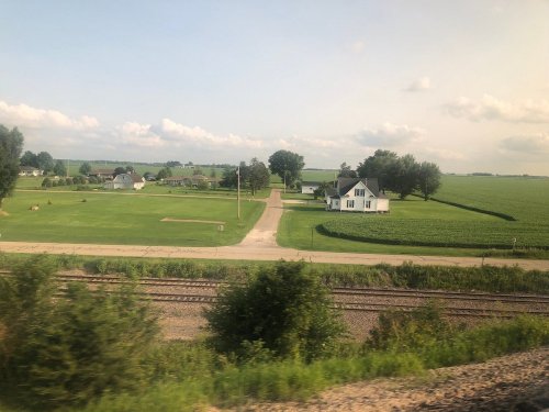 A rural landscape featuring a white house, green fields, and a dirt road alongside train tracks under a cloudy sky.