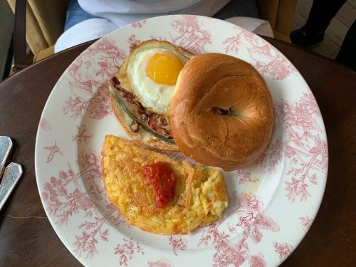 A plate with a bagel sandwich topped with an egg, alongside a potato hash with a tomato slice.