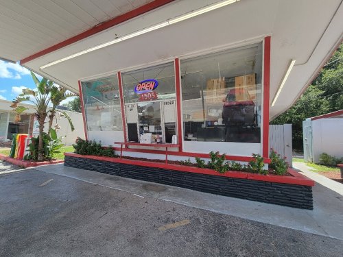A small storefront with large windows, an "OPEN" sign, and a flower bed in front, set against a clear blue sky.