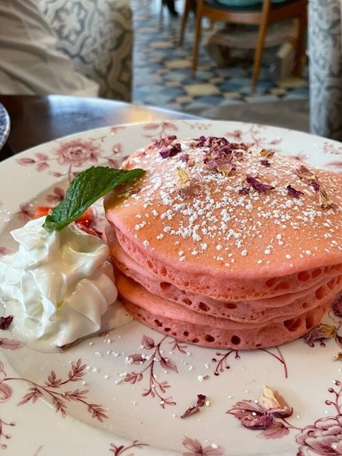 Three pink pancakes topped with powdered sugar, rose petals, and a mint leaf, served with a dollop of whipped cream.