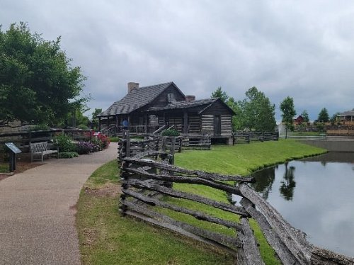 A rustic log cabin by a pond, surrounded by greenery and a wooden fence, under a cloudy sky.