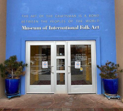 Entrance to the Museum of International Folk Art, featuring a blue wall and double glass doors with decorative planters.
