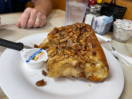 A large slice of pecan pie on a plate, accompanied by a small container of cream, with a glass of water in the background.